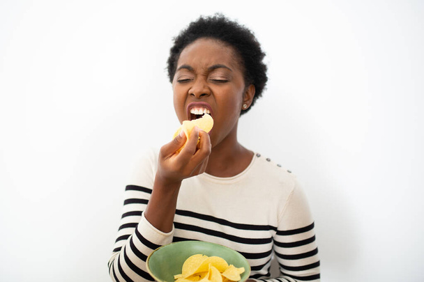 Portrait hungry African American woman eating chips by isolated white background  - Photo, image