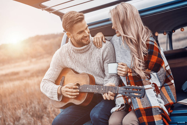Handsome young man playing guitar for his girlfriend while both sitting in the car trunk outdoors - Foto, imagen