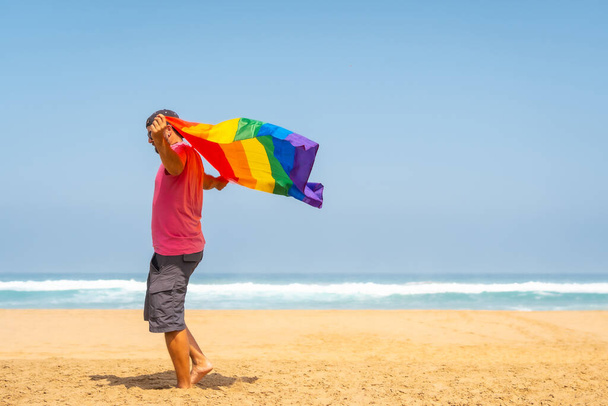 A gay person with a pink t-shirt and black cap with the LGBT flag on a beach, symbol of homosexuality - Photo, image