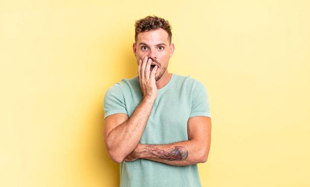 young handsome hispanic man open-mouthed in shock and disbelief, with hand on cheek and arm crossed, feeling stupefied and amazed - Fotoğraf, Görsel
