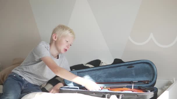 Το παιδί δεν θέλει να κάνει βιολί στο σπίτι, το αγόρι κάθεται στο κρεβάτι δίπλα στο ανοιχτό εξώφυλλο με το βιολί. - Πλάνα, βίντεο