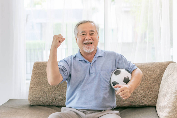 Возбужденный пожилой человек, старик чувствовать себя счастливым держа футбольный мяч готовиться к команде чирлидеров фаворит на диване дома, образ жизни Азиатский Старший хорошая концепция здоровья - Фото, изображение