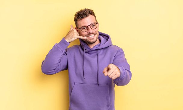 young handsome hispanic man smiling cheerfully and pointing to camera while making a call you later gesture, talking on phone - Fotografie, Obrázek