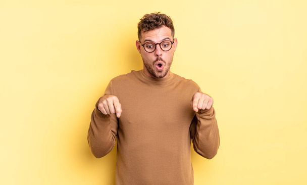 young handsome hispanic man with open mouth pointing downwards with both hands, looking shocked, amazed and surprised - Foto, afbeelding
