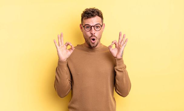 young handsome hispanic man feeling shocked, amazed and surprised, showing approval making okay sign with both hands - Fotoğraf, Görsel