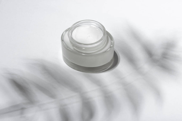 Cosmetic cream or mask in a glass jar with hard shadows from the leaves on a white background. Natural organic care for face and body skin. View from above. - Φωτογραφία, εικόνα