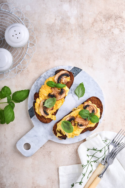Scrambled eggs with fried mushrooms and basil on bread on white table background. Homemade breakfast or brunch meal. Scrambled eggs and mushrooms sandwiches. Top view with copy space - Zdjęcie, obraz