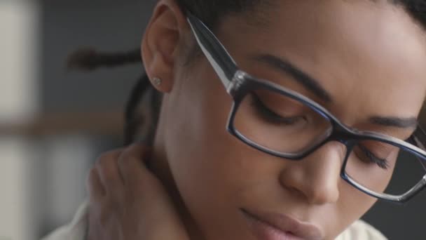 Nuori järkyttynyt afrikkalainen amerikkalainen nainen silmälaseissa, joka kärsii kaulasta, hieroo lihaksiaan toimistossa, lähikuva - Materiaali, video