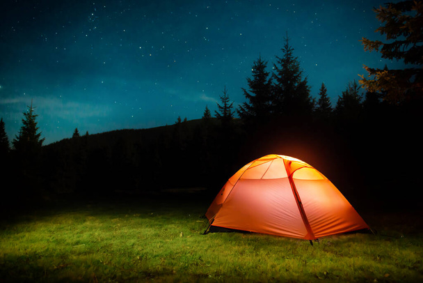 Tienda iluminada naranja en bosque nocturno oscuro con cielo nocturno y estrellas - Foto, imagen