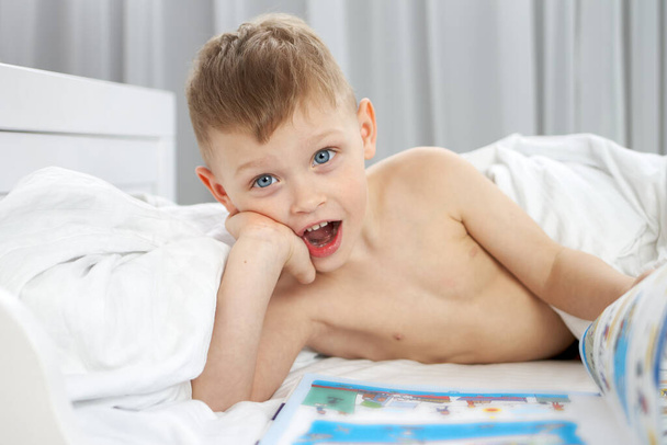 The boy smiles as he looks at the picture book covered with a white blanket - Foto, imagen