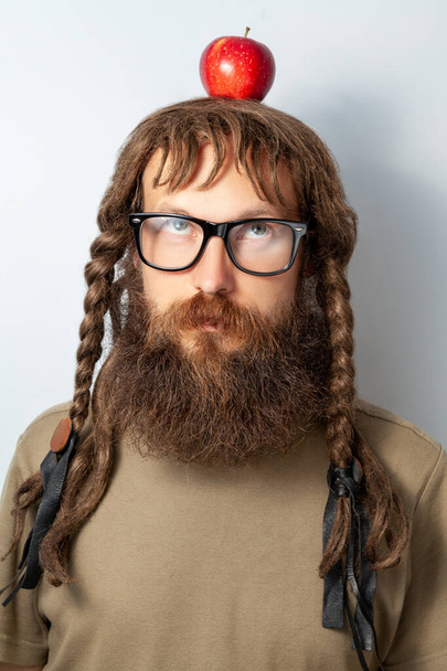 Handsome male scientist or professor with long beard and pigtails holding apple on head, newtons law, bald bearded man wearing T-shirt and glasses. Indoor studio shot isolated on gray background. - Φωτογραφία, εικόνα