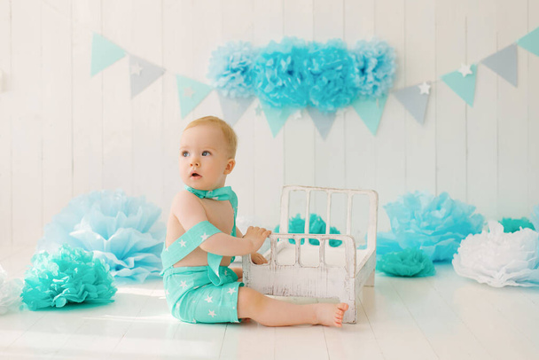 Egy egyéves kisfiú egy nyári öltönyben pillangóval ül a születésnapja fotózónáján kék színekben egy babaággyal. - Fotó, kép