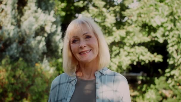 Όμορφη χαμογελαστή ηλικιωμένη γυναίκα στέκεται στο καλοκαιρινό κήπο και κοιτάζοντας κάμερα, - Πλάνα, βίντεο