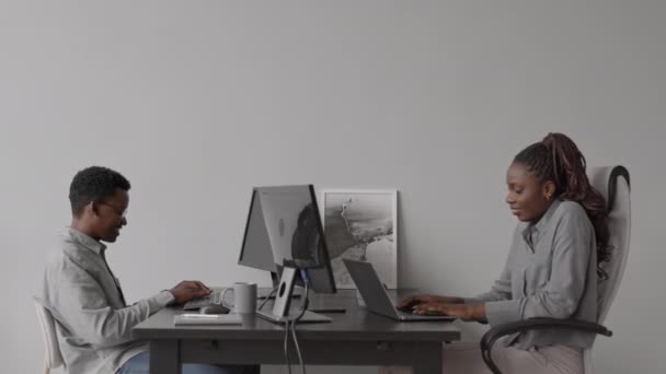 Side-view közepes szúrás lövés két afro-amerikai informatikai programozó ül asztaloknál egymás előtt a modern irodában dolgozik PC-k - Felvétel, videó