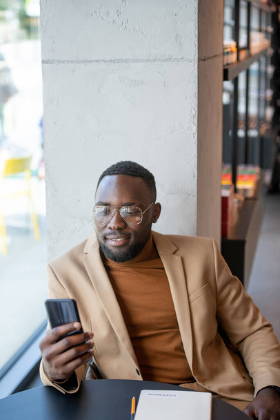 African man scrolling in smartphone by table in bookshop - Photo, Image
