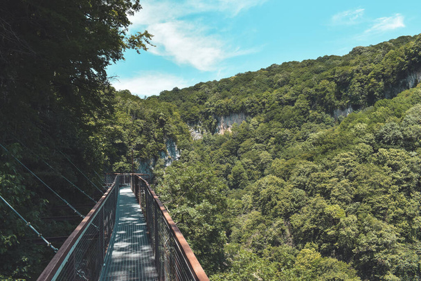 Puente colgante en Okatse Canyon, aldea Gordi, región de Imereti, Georgia. Sendero estrecho rodeado de colinas cubiertas de árboles verdes. Cielo azul con nubes claras arriba. Atracción turística georgiana - Foto, Imagen