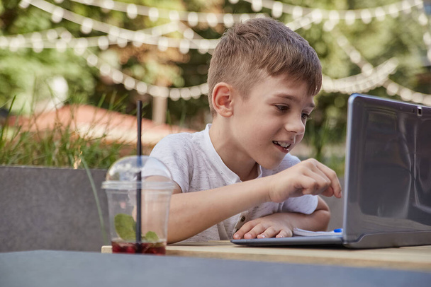 sorrindo menino caucasiano sentado à mesa na varanda do café, estudando com laptop - Foto, Imagem