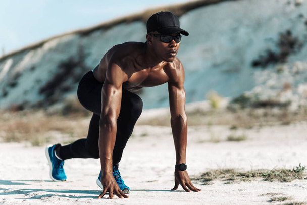 Apto e confiante afro-americano homem, na posição inicial pronto para correr, atleta masculino prestes a começar um sprint, ao ar livre na natureza, com luz solar brilhante, conceito de esporte, início, motivação - Foto, Imagem
