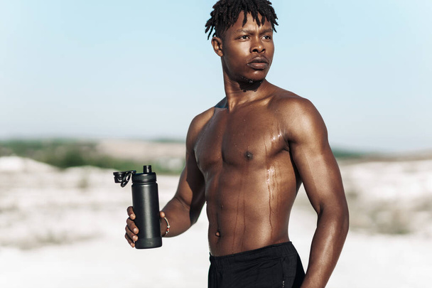 Portrait African American man, fitness athlete, with a bottle of water after training on a sunny day outdoors in nature, sports, motivation, healthy lifestyle - Valokuva, kuva