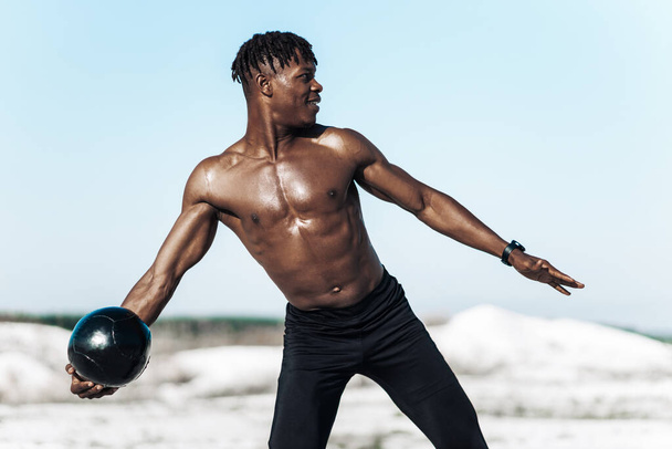 African American crossfit man, holding ball over head for shoulder press workout, outdoors against mountains background, muscular athlete doing upper body workout working out with heavy weighted balls - Фото, изображение
