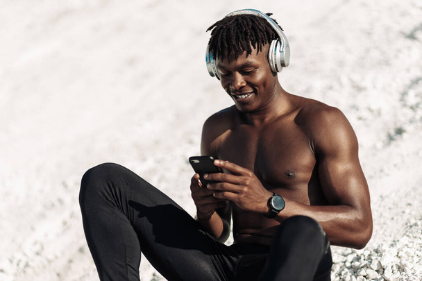 Smiling sporty African American man using mobile phone and enjoying music, athlete goes in for sports outdoors - Valokuva, kuva