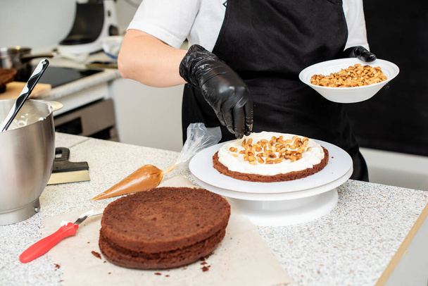 Konditor oder Bäcker in schwarzen Handschuhen und schwarzer Küchenschürze backen einen Kuchen. Hausgemachte Geburtstagstorte. Konzept von hausgemachtem Gebäck, Kuchen kochen, Hobby, weibliche Kleinunternehmen - Foto, Bild