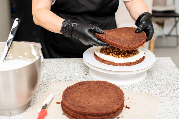 Ζαχαροπλαστική σεφ μάγειρας ζαχαροπλάστης ή αρτοποιός σε μαύρα γάντια και μαύρη ποδιά κουζίνας κάνει μια τούρτα. Σπιτική τούρτα γενεθλίων. Έννοια της σπιτικής ζαχαροπλαστικής, κέικ μαγειρικής, χόμπι, γυναικεία μικρή οικιακή επιχείρηση - Φωτογραφία, εικόνα