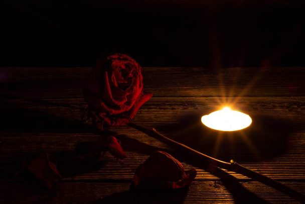 Una rosa roja seca, pétalos secos junto a una vela de té sobre una mesa de madera sobre un fondo negro y oscuro. Dramática atmósfera mística ritual de magia negra. El concepto de duelo y duelo postal. - Foto, imagen