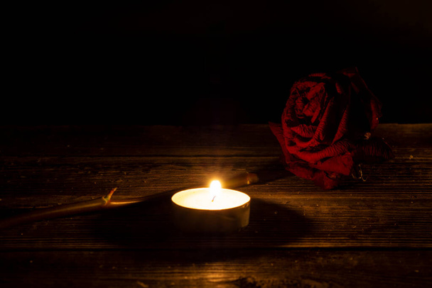 Jedna czerwona suszona róża, suche płatki obok świecy herbaty na drewnianym stole na czarnym, ciemnym tle. Dramatyczna mistyczna atmosfera, rytuał czarnej magii. Pojęcie żałoby i żałobnej pocztówki. - Zdjęcie, obraz