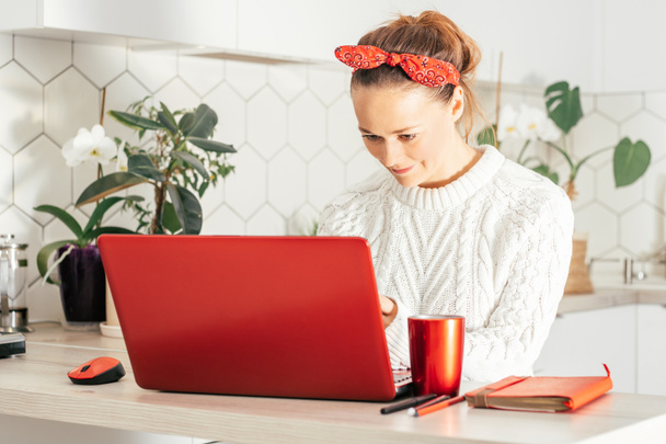 Young woman sits with red laptop at home in kitchen in red bandage and white sweater and looks at monitor. Home work or study concept - Valokuva, kuva