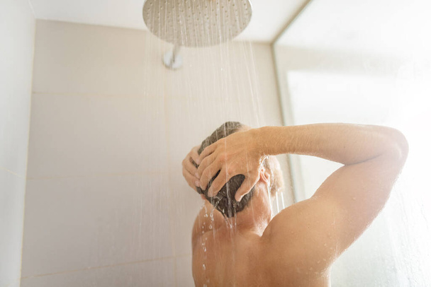 Man taking a shower washing hair under water falling from rain showerhead in luxury walk-in bath. Showering young person at home lifestyle. Body care morning routine in sunlight - Fotoğraf, Görsel
