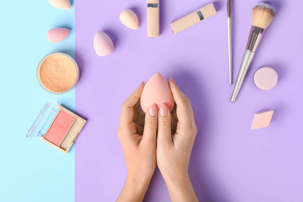 Жіночі руки зі стильною губкою для макіяжу та декоративною косметикою на кольоровому фоні
 - Фото, зображення