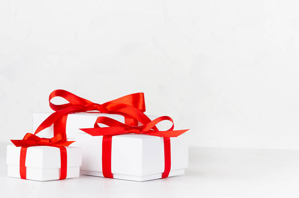Three festive white paper boxes for gift with red silk ribbon and bow on white table in elegant minimal style, copy space. - Foto, Imagem
