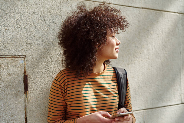 Cute black girl looking away while holding a cellphone outdoors during the day - Foto, Bild