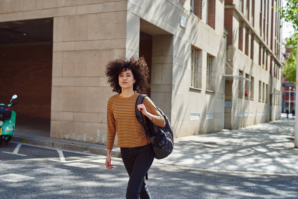 Attractive black girl crossing a street while walking with a backpack outdoors during the day - 写真・画像