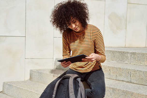 Beautiful black girl reading from her tablet pc while sitting with a backpack on a flight of stairs outdoors - Foto, Bild