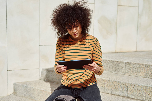 Cute black girl reading something on her tablet pc while sitting alone on a flight of stairs outdoors during the day - Foto, Bild
