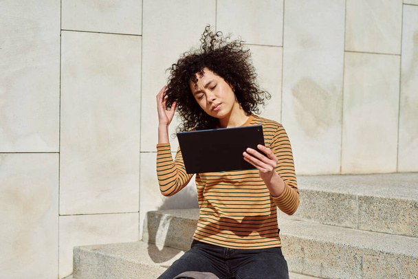 Nettes schwarzes Mädchen liest etwas auf ihrem digitalen Tablet, während es an einem sonnigen Tag allein auf einer Treppe im Freien sitzt - Foto, Bild