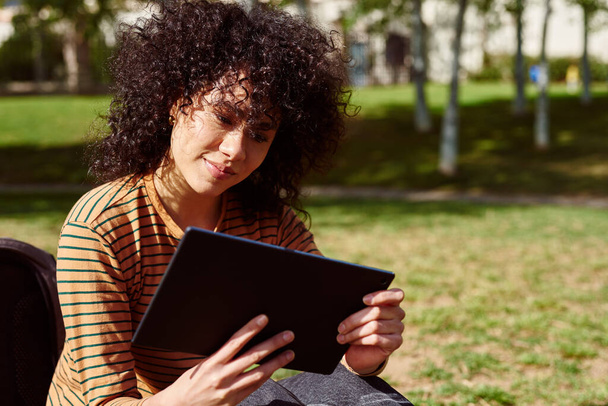 Attractive young black woman reading from her tablet pc while sitting alone in a park during the day - Photo, Image