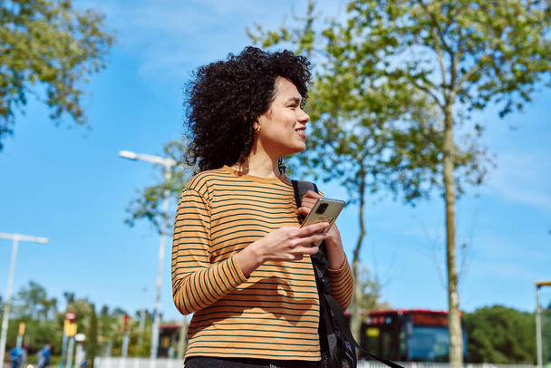 Linda mujer negra joven mirando hacia otro lado alegremente mientras sostiene un teléfono celular al aire libre durante el día - Foto, Imagen