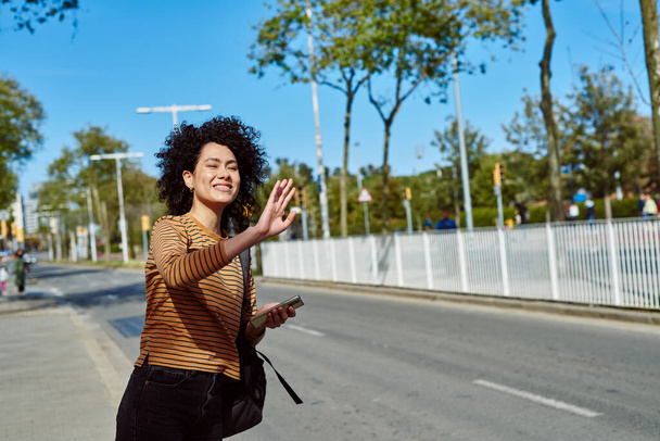 Happy young black woman hailing a taxi while standing by the roadside with a cellphone and a backpack during the day - Foto, afbeelding
