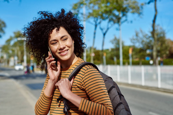 Happy young black woman taking a phone call while walking with a backpack outdoors on a sunny day - 写真・画像