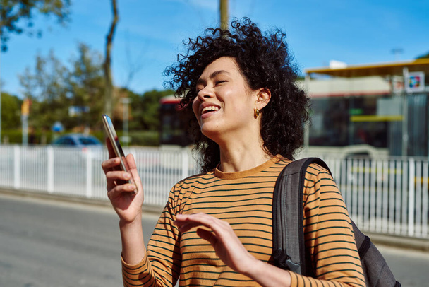 Laughing young black woman holding a cellphone while standing with a backpack by the roadside during the day - Photo, Image