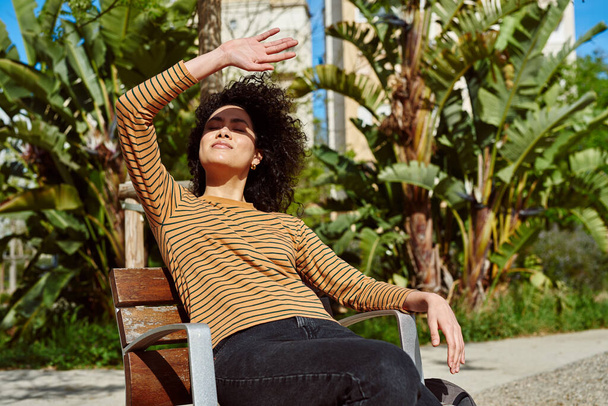 Attractive young black woman blocking the sun from her eyes while relaxing on a chair outdoors during the day - Foto, Imagem