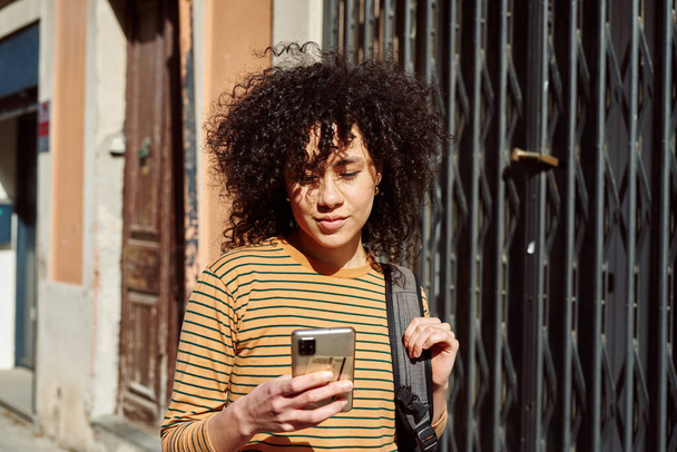Aranyos fiatal fekete nő használja a mobiltelefon séta közben egy hátizsák a szabadban a nap folyamán - Fotó, kép