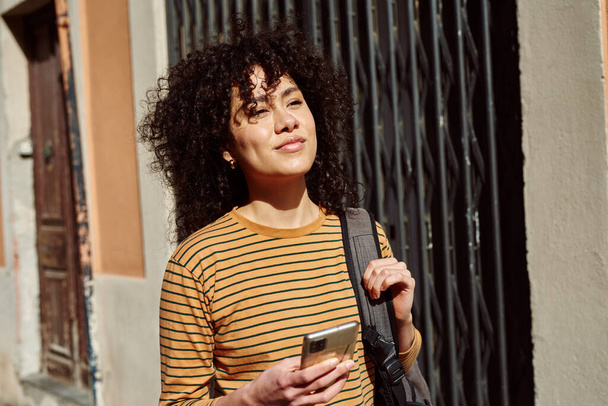 Cute young black woman looking thoughtful while holding a cellphone and carrying a backpack outdoors during the day - 写真・画像