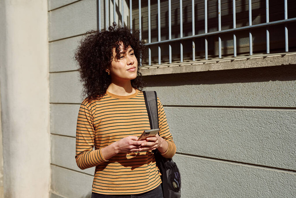 Cute young black woman looking away thoughtfully while holding a cellphone and carrying a backpack outdoors during the day - 写真・画像