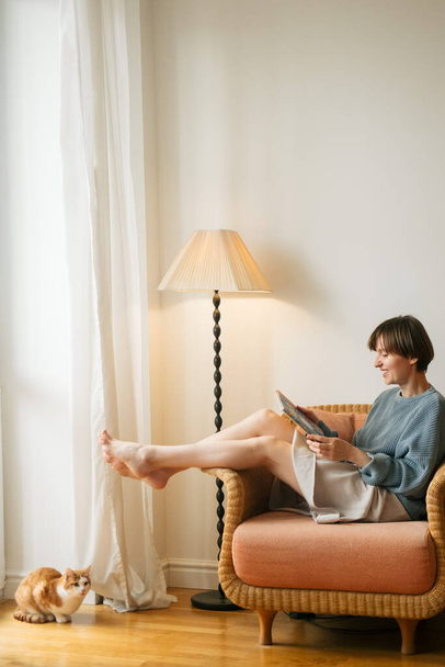 Beautiful young relaxed woman is sitting in chair in bedroom and reading book. Cozy home weekends and reading hobby concept. - Valokuva, kuva