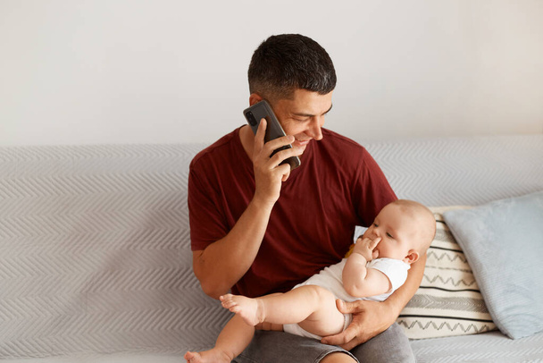 Side view portrait of positive dark haired father talking via cell phone and holding infant daughter in his hands, having pleasant conversation, spending time with his child. - Fotografie, Obrázek