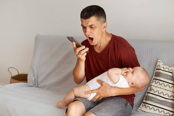 Wściekły nieszczęśliwy ojciec w burgundowym stylu t-shirt siedzi na kanapie z córkami, trzymając smartfon, krzycząc z gniewu, wyrażając agresję. - Zdjęcie, obraz